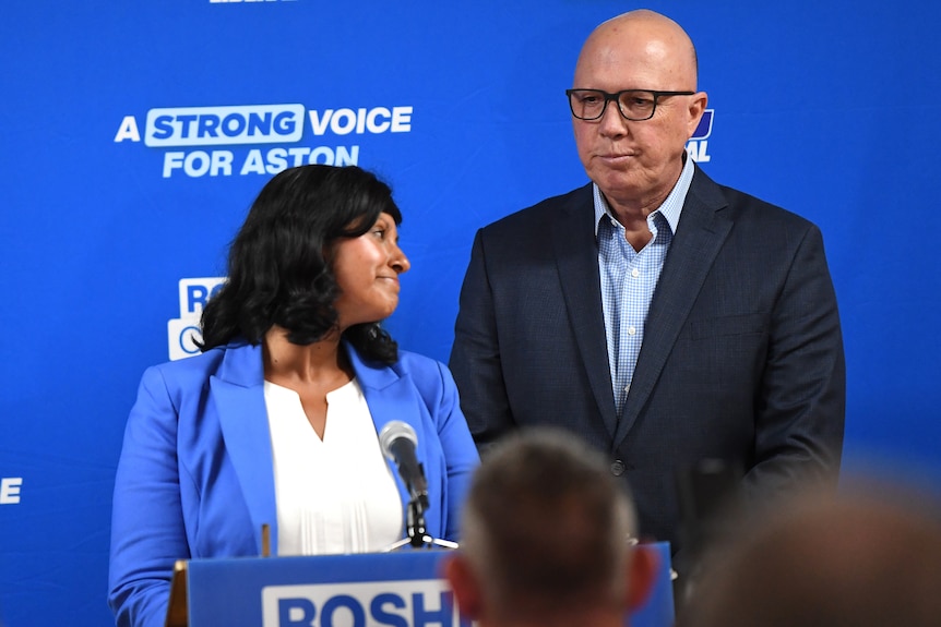 Roshena Campbell and Peter Dutton look downcast while standing on a blue stage.