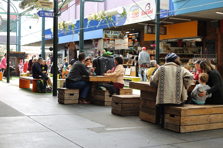People sit at wooden benches eating food and talking inside Preston Market.