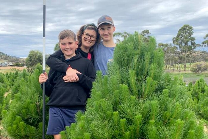 A mother and two sons hugging and smiling, behind a Christmas tree
