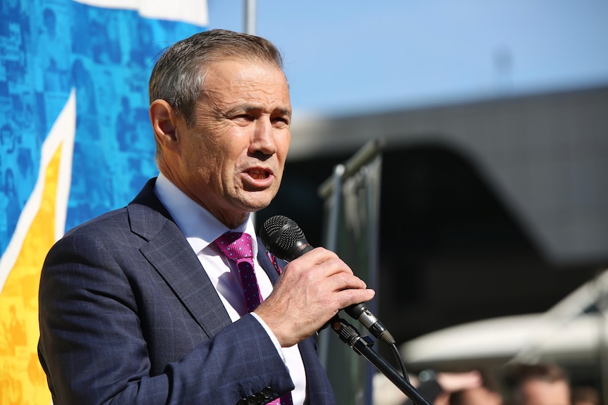 A close up of Roger Cook wearing a blue suit and pink tie, holding a microphone on a podium at a rally.