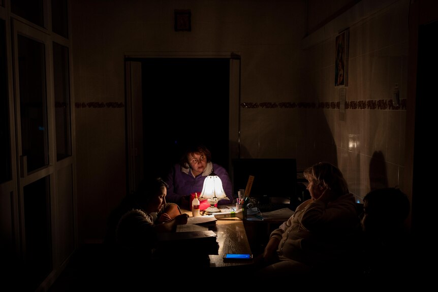 Three women sit around a table with a lamp lighting up their faces. 