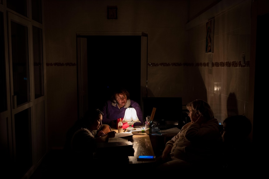 Three women sit around a table with a lamp lighting up their faces. 