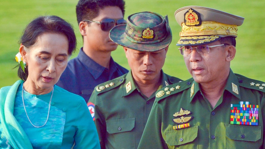 Aung San Suu Kyi in abito blu che cammina con due uomini in uniforme dell'esercito