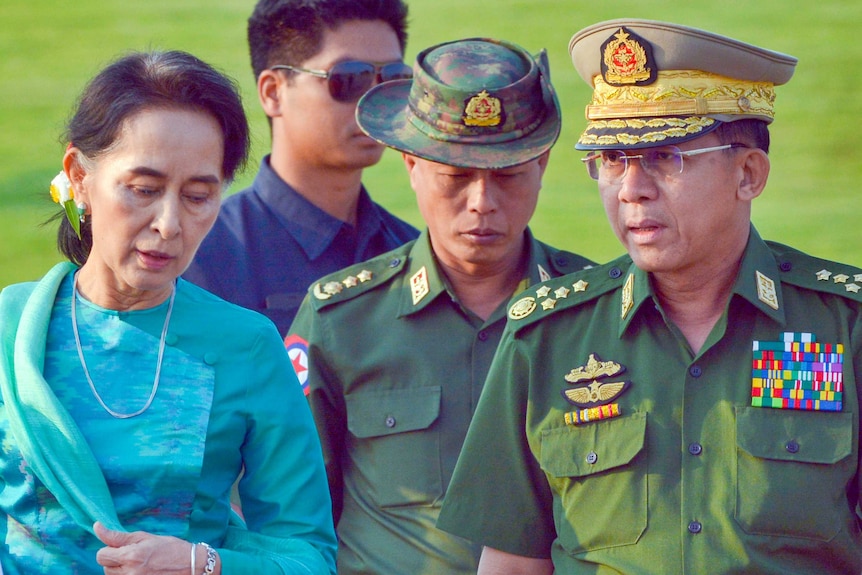 Aung San Suu Kyi in a blue dress walking with two men in army uniforms