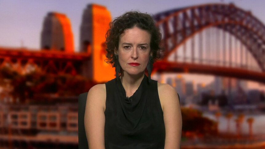 Woman with black curly hair, wearing black sleeveless top, looks at the camera. Sydney Harbour Bridge backdrop.