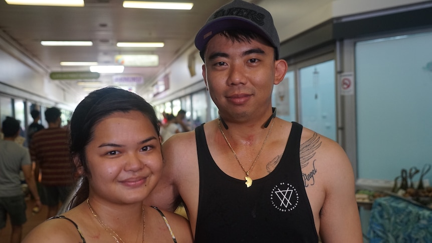 Angel and Randy Ku are wearing singlets and smiling slightly to the camera. A busy market is behind them.