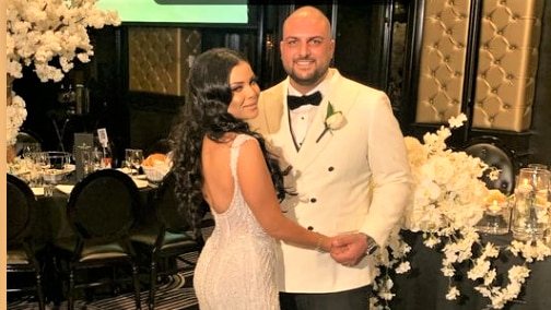 A bride and groom at their wedding reception. The bride has a backless dress and long dark hair. The groom has a white coat.