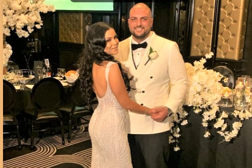 A bride and groom at their wedding reception. The bride has a backless dress and long dark hair. The groom has a white coat.