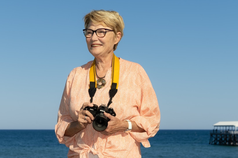 Woman with blonde hair and glasses holding SLR camera with strap around neck looking to the left at beach on a sunny day.