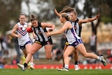 Four AFLW players converge in the centre, with the woman second from left holding the football.