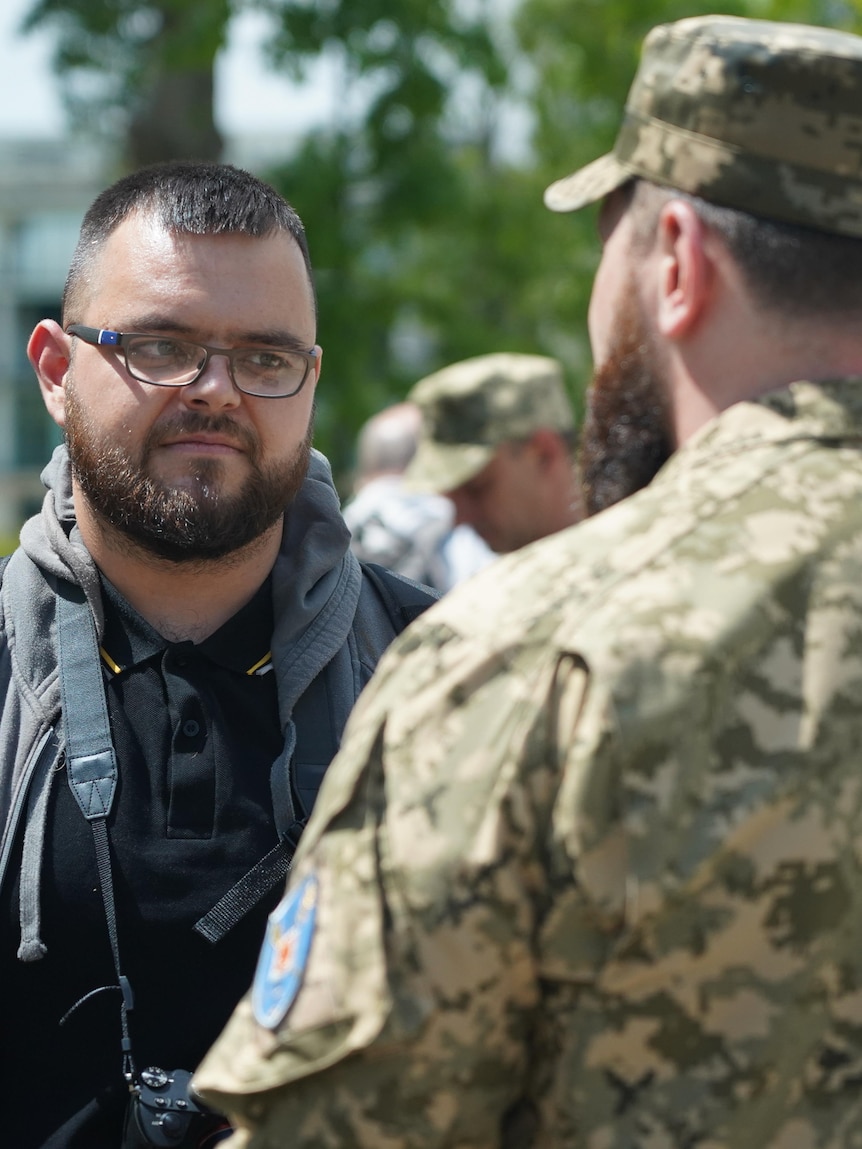 A man with close-cropped hair and beard, and glasses, standing in front of a man in army fatigues.