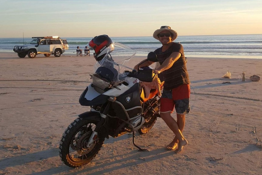 A smiling Jaybin Henry stands next to a BMW motorbike on Cable Beach in Broome with a 4WD in the background.