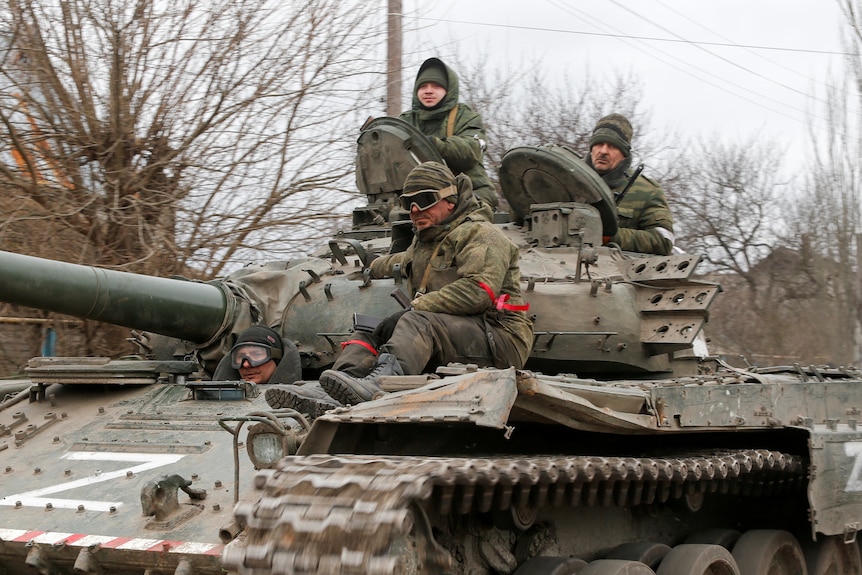 dark green uniforms ride on top of a tank, they are wearing white and red armbands.Men in 