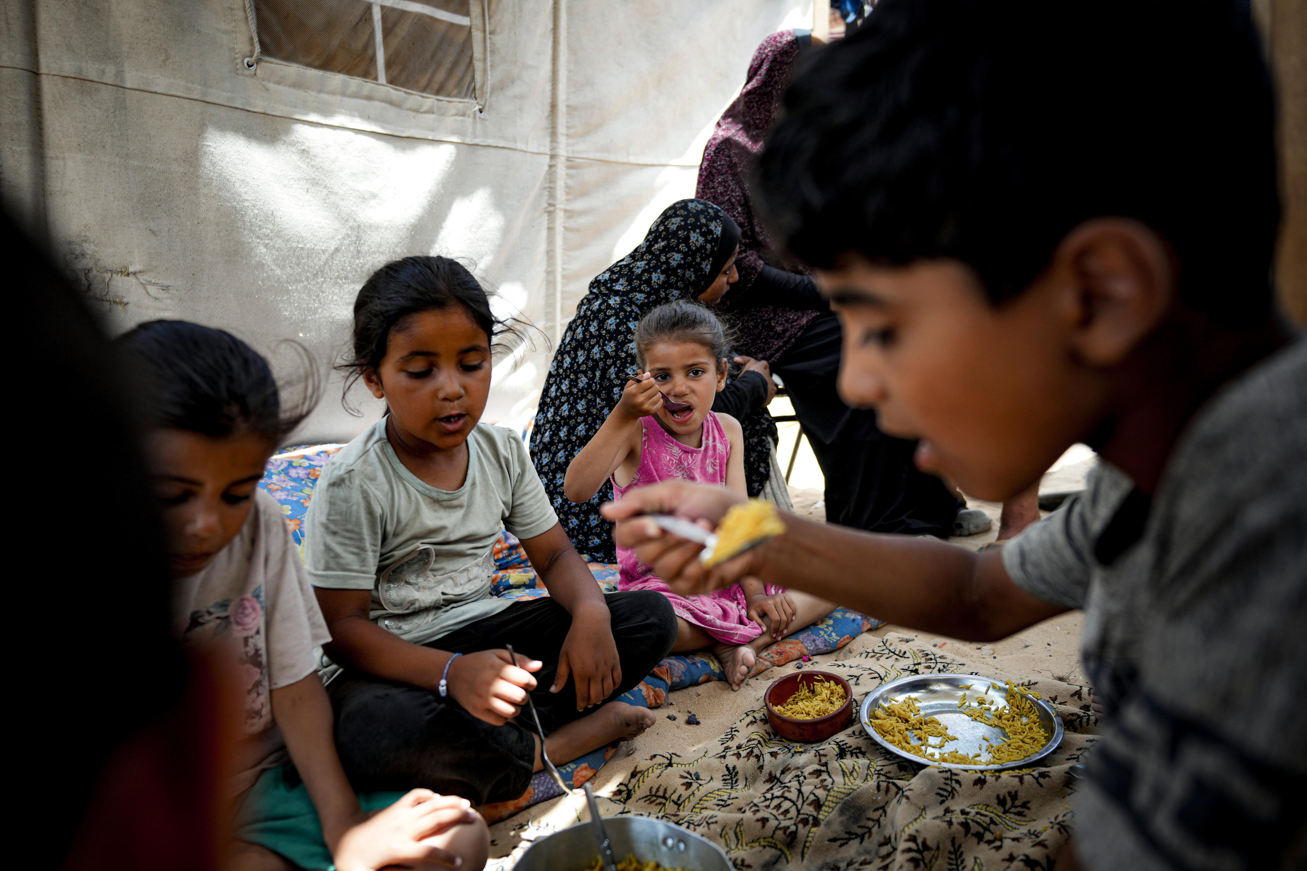 Four young children eat rice sitting on a blanket on the floor, two women are behind them