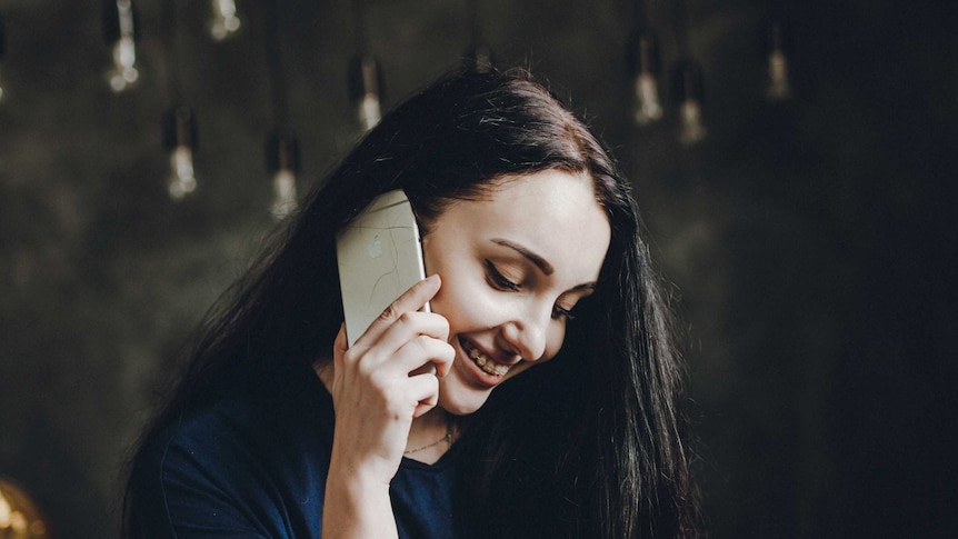 Woman with brown hair holds white iphone to her hear on a phone call, she is smiling