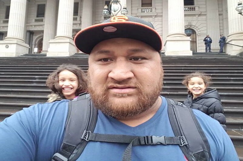 Clarence Leo posing for a selfie with two kids in the background on the steps of the Victorian Parliament.