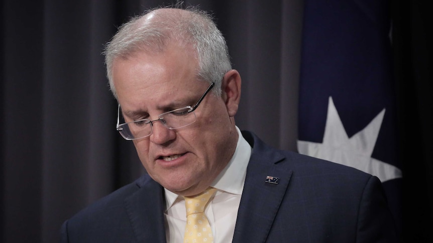 Morrison is looking downwards, a bit grim, wearing a blue suit, yellow tie and Australian flag pin.