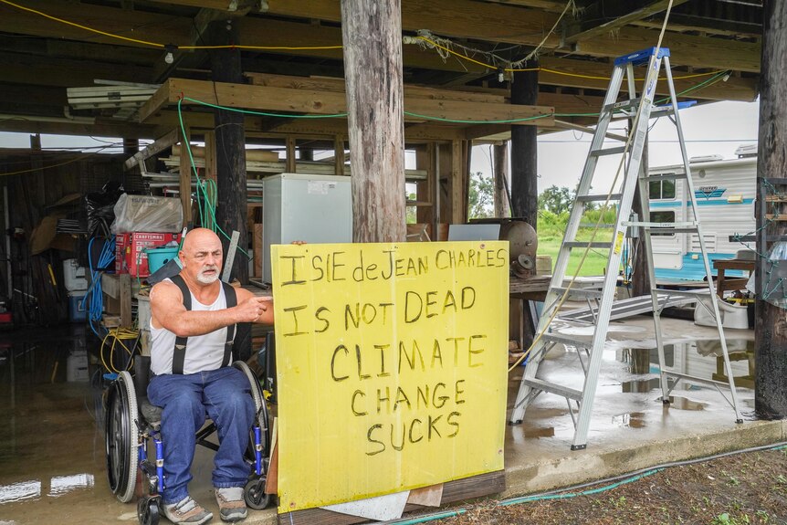 Chris Brunet in a wheelchair next to a sign that reads The Isle of Jean Charles is not dead Climate change stinks