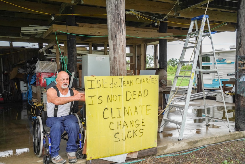 Chris Brunet na wózku inwalidzkim obok tabliczki z napisem, że Wyspa Jean Charles nie umarła Zmiany klimatyczne śmierdzą