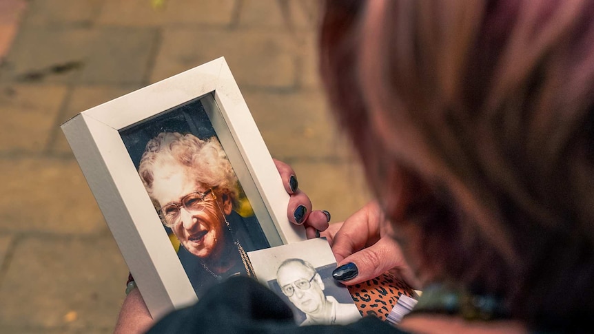A woman holds a framed picture of an older woman with white hair wearing glasses, smiling.