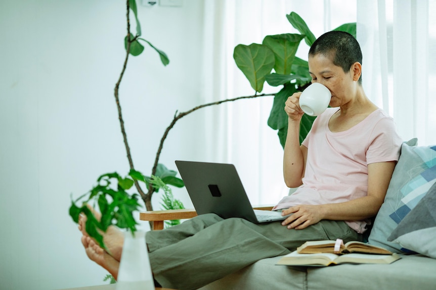 A woman with a shaved head sits on the couch drinking from a mug and looking at a laptop.
