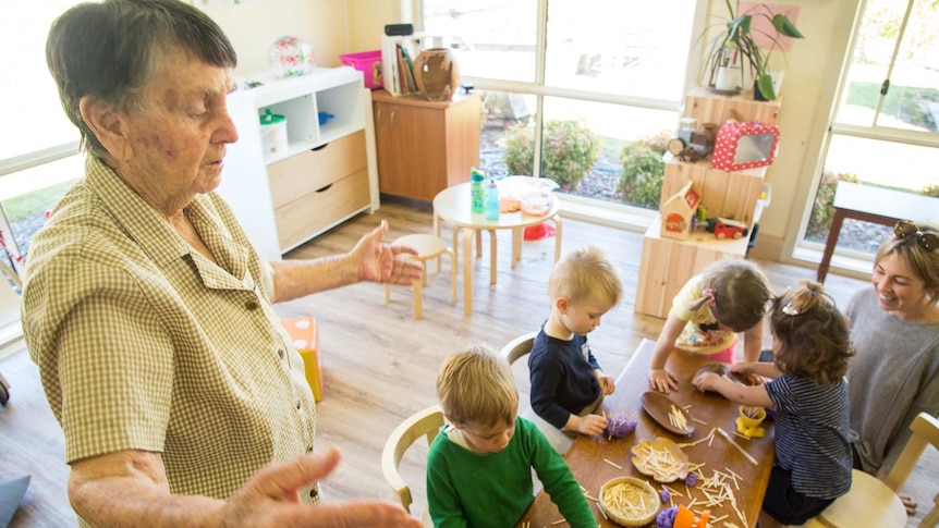 An elderly resident with dementia hovers near a low table where children are having fun with playdough.