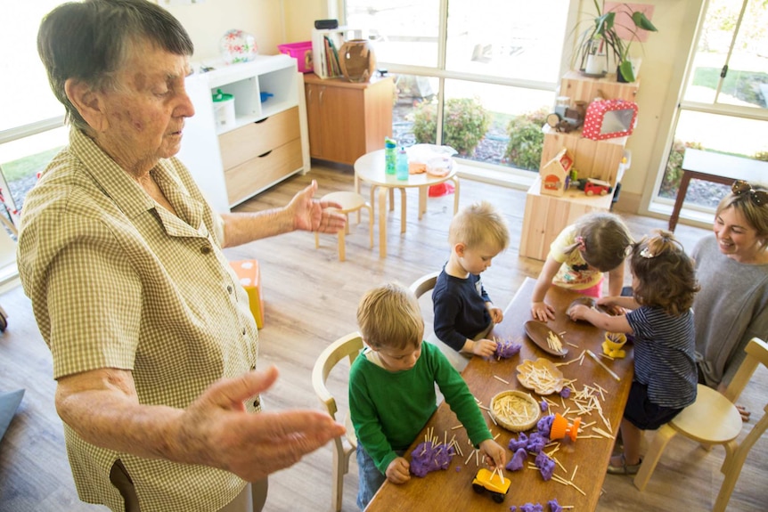 An elderly resident with dementia hovers near a low table where children are having fun with playdough.