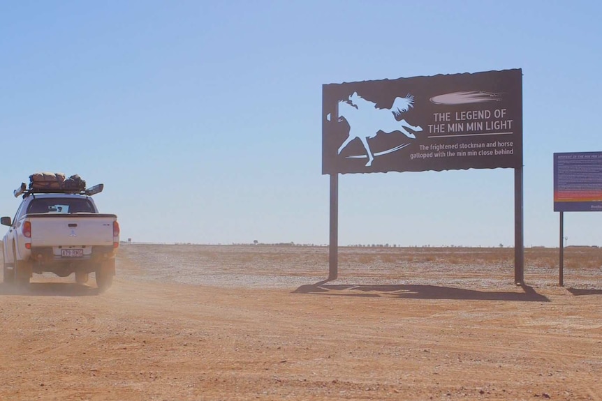 A white ute travelling on a red dirt road turns at a sign which reads 'the legend of the min min light'.