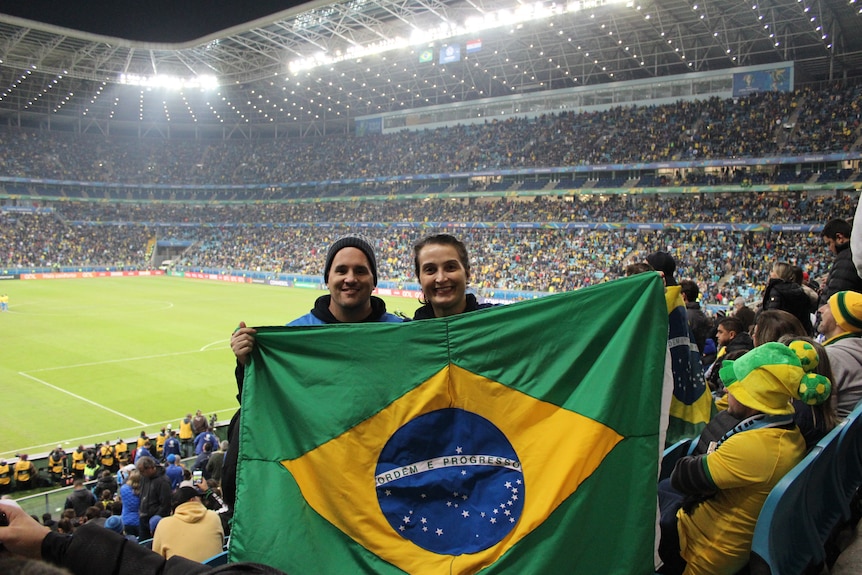 A young man and woman hold up a Brazilian flag in a crowded soccer stadium