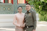 A young Thai woman in a traditional chut thai, and a man in a green police uniform