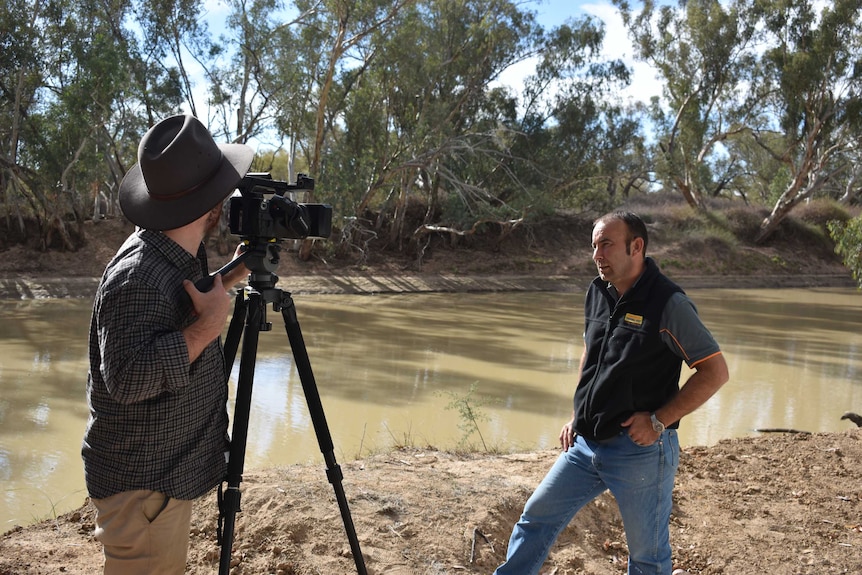 A man wearing an Akubra standing behind a camera, filming another man by a river.