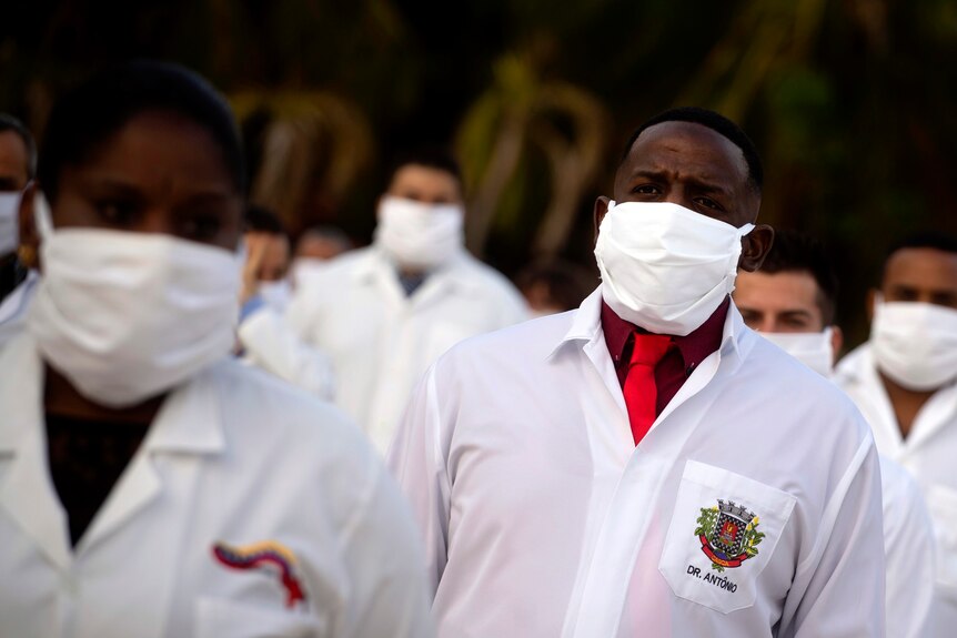 Doctors and nurses wearing medical coats and surgical masks stand in rows, with their hands behind their backs.