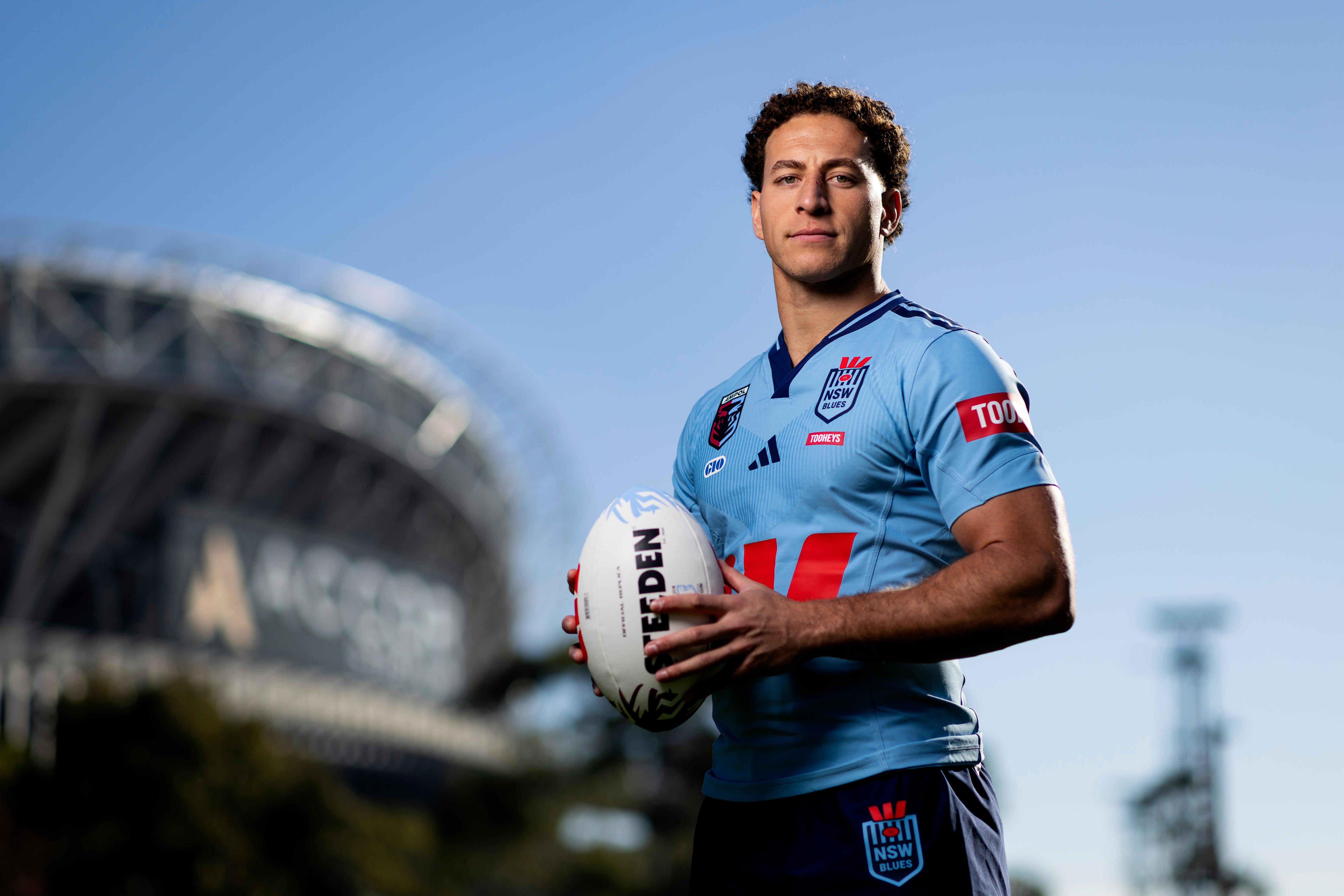 Rugby league player posing for a portrait while holding a football. A stadium is in the background to the left.