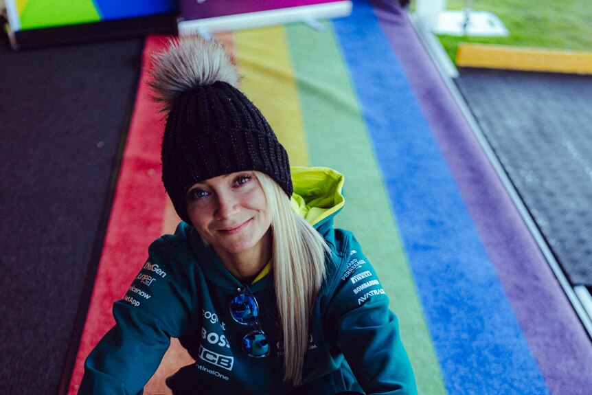A young woman wearing a beanie and jacket sits on a rainbow flag mat. 