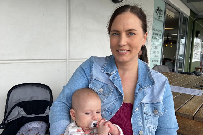 A woman in a blue denim jacket sitting on a bench holding a baby