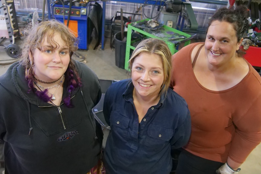 Three women in a metalworking workshop, looking up to camera and smiling.