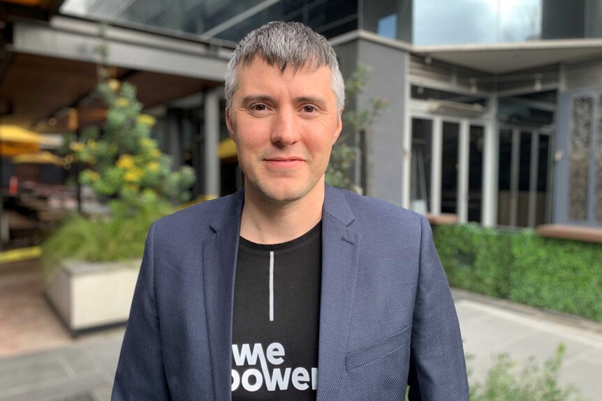 Kaspar Kaarlep in a "WePower" t-shirt and grey blazer stands in front of a modern building.