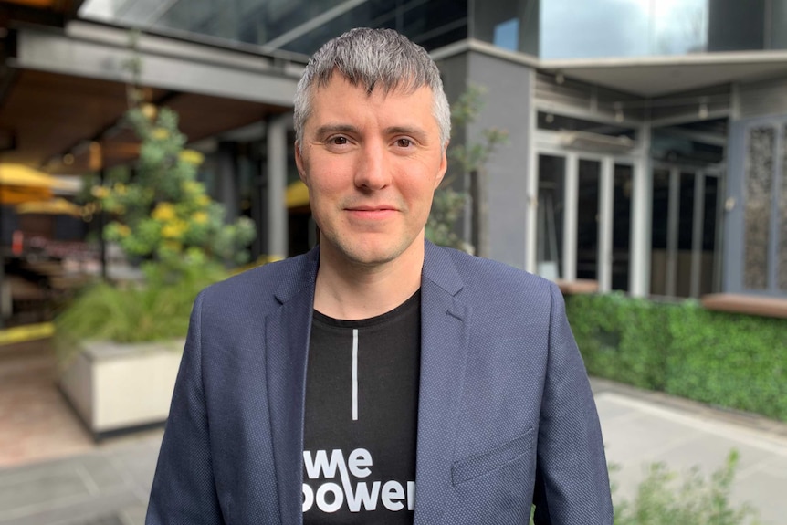 Kaspar Kaarlep in a "WePower" t-shirt and grey blazer stands in front of a modern building.