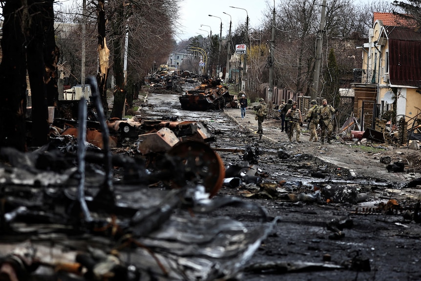 Soldiers walk past a destroyed Russian tank and armoured vehicles on a deserted street.