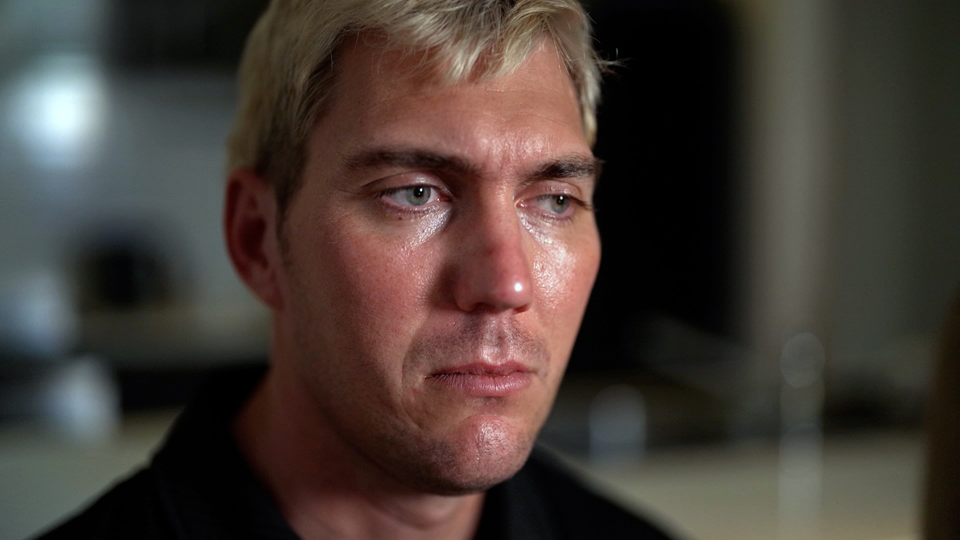 A close-up of a man sitting indoors, looking ahead. His expression is sad.