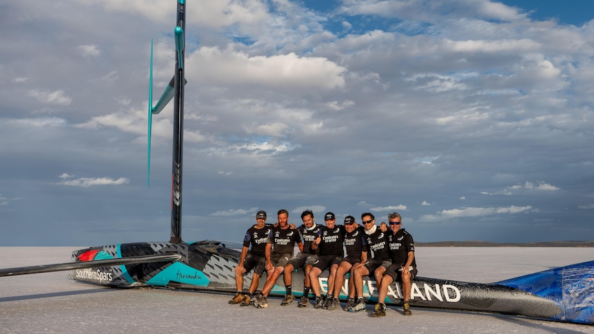 A group of men dressed in black sitting on a fibreglass aerodynamic vehicle on a salt lake
