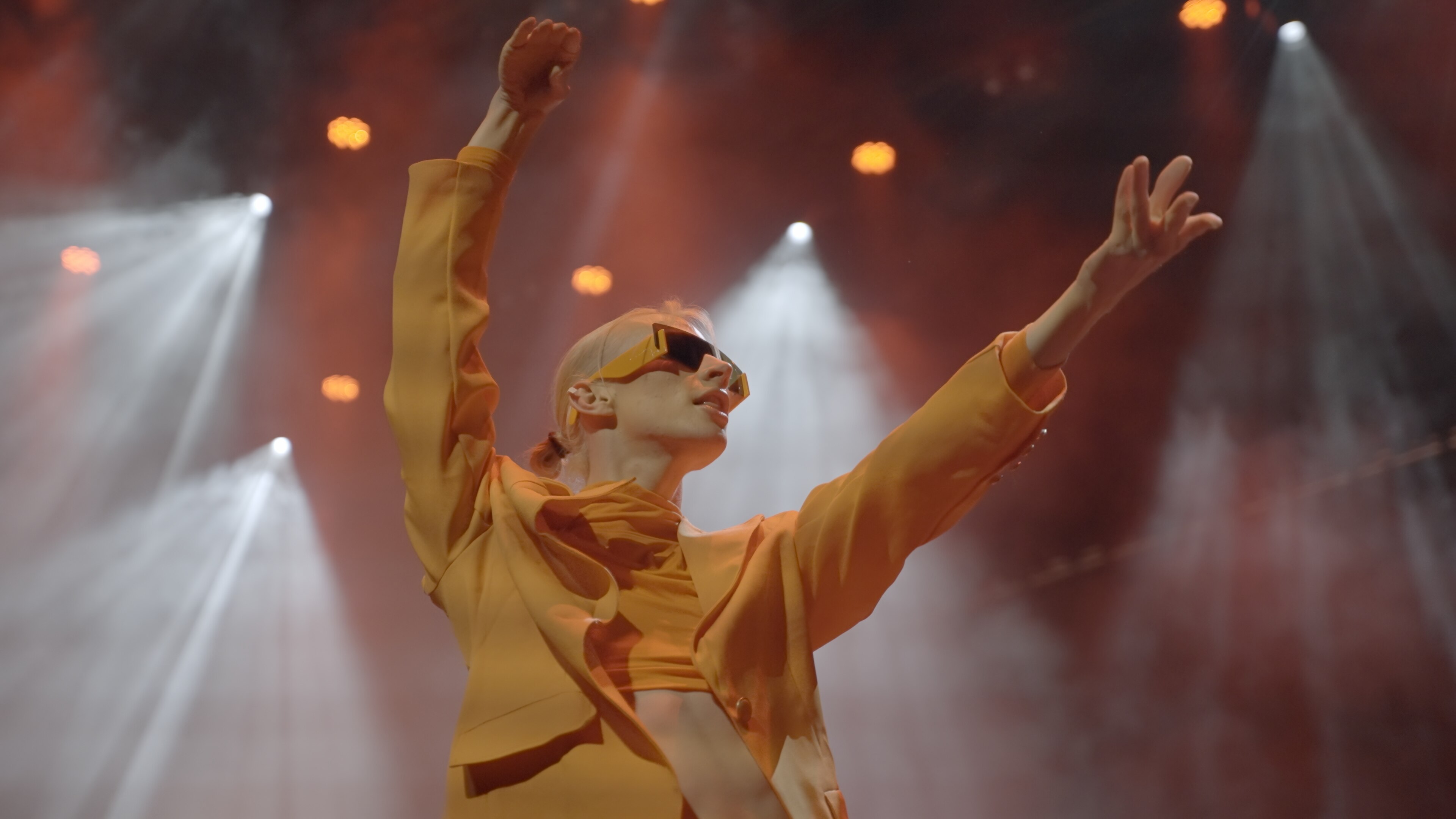 A slim dancer with blonde hair wearing sunglasses and a yellow top flings their arms out on stage