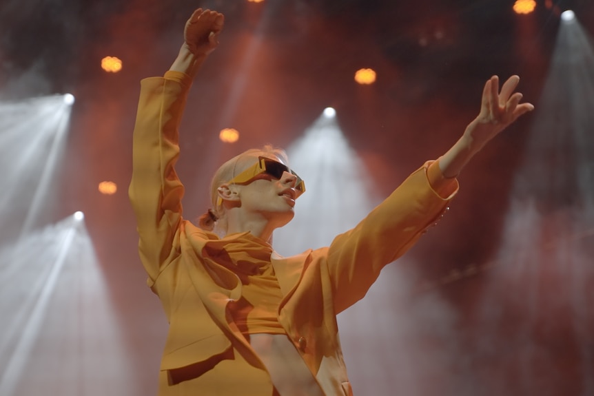 A slim dancer with blonde hair wearing sunglasses and a yellow top flings their arms out on stage