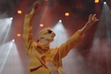 A slim dancer with blonde hair wearing sunglasses and a yellow top flings their arms out on stage