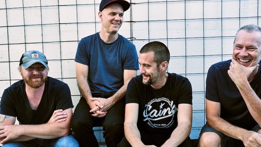 four men sit side by side wearing t shirts and smiling
