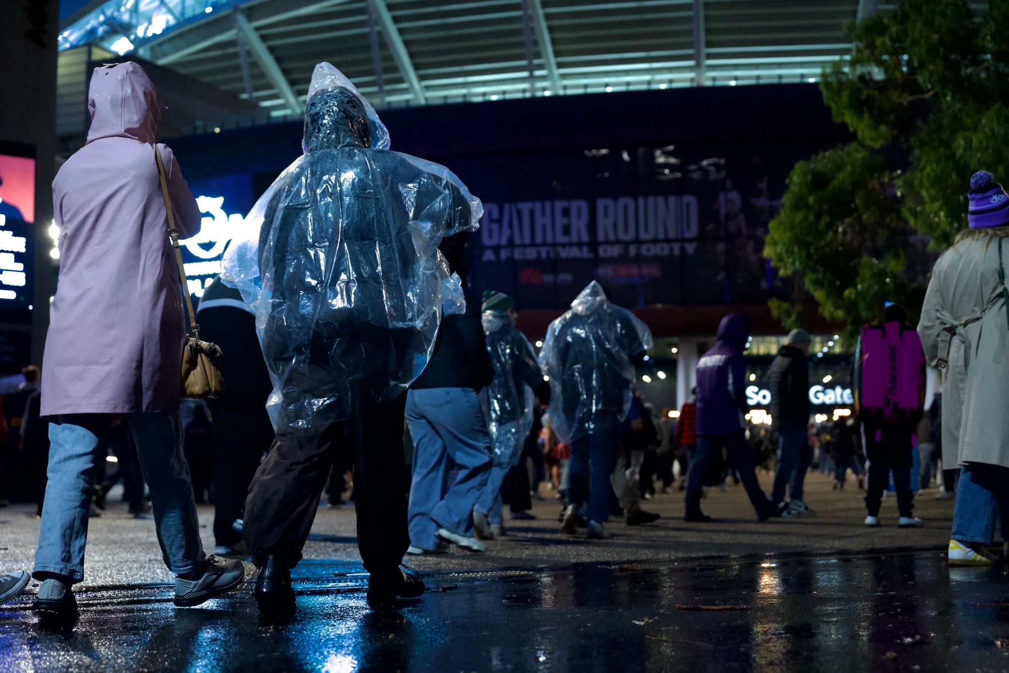 Pessoas com capas de chuva ou capuzes enquanto caminham em direção a um estádio esportivo onde telas externas exibem cartazes do Gather Round