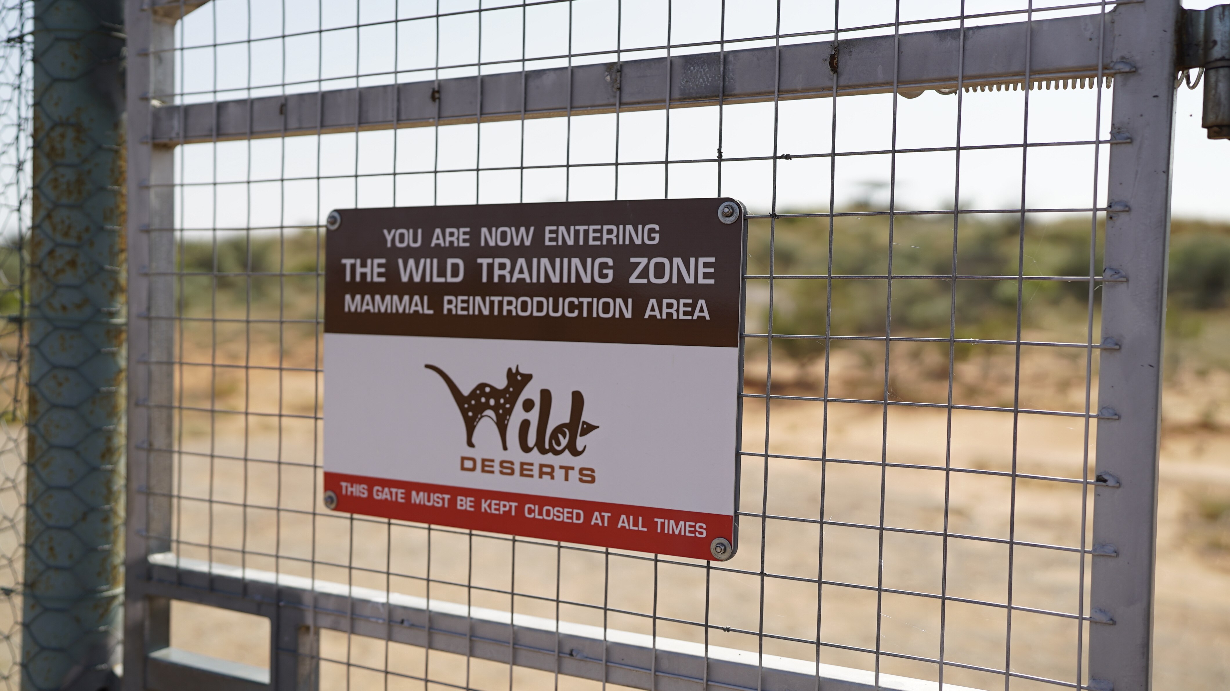 A sign for the Wild Deserts mammal reintroduction training zone on a metal fence.