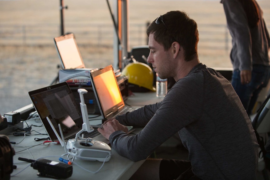 A man sits at a desk at an airport with a drone controller on the desk