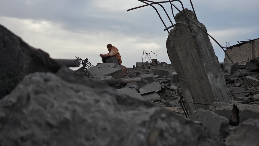 A man sits on top a pile of concrete rubble. In the foreground, twisted metal sticks out of a chunk of concrete