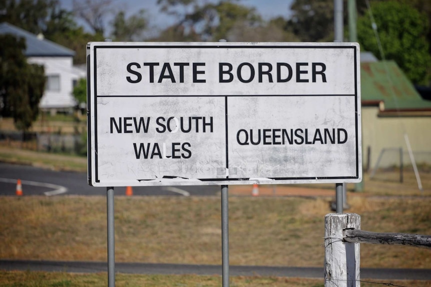 A decaying and dirty white sign saying 'state border, NSW and Queensland' with houses and trees behind.